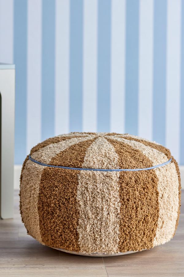 Charoline Pouf Brown – Bloomingville MINI : pouf devant un mur habillé de papier peint à rayures bleues.